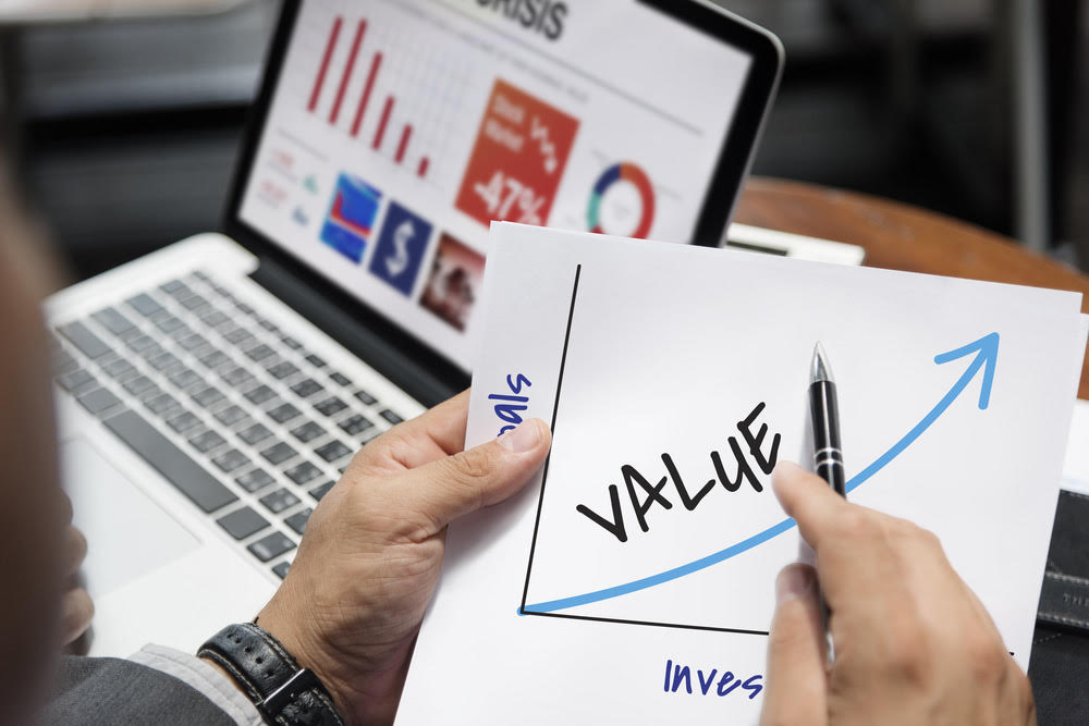 Rule one investing vs value investing: A hand holds up a chart with value going up, a computer with stock diagrams is also in the background