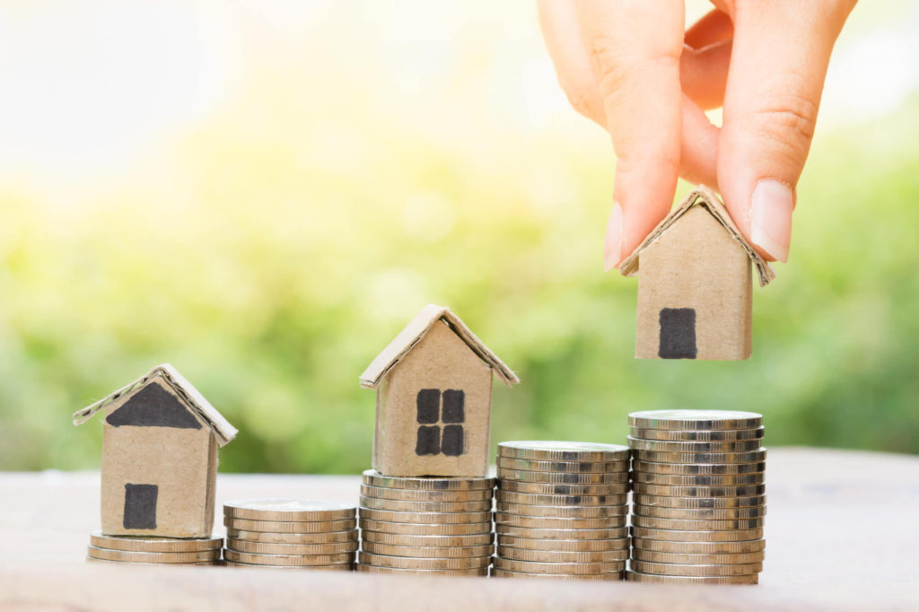 Investment ideas: three cardboard houses stacked on coins going upwards. The last house is being placed on by fingers.