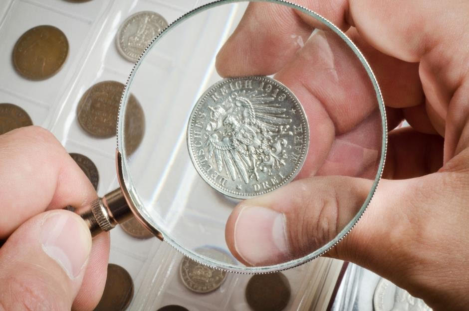 Collect coins: two hands inspect a 1918 German coin using a magnifying glass with the rest of their coin collection in the background