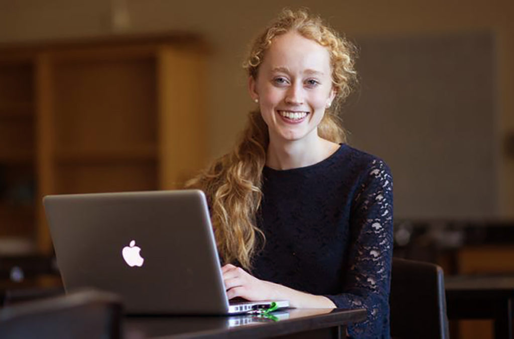 Erin Smith: a young woman sits smiling at the camera with an Apple MacBook in front of her