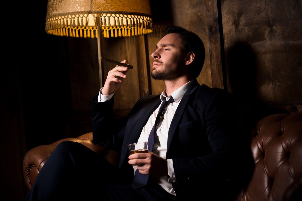 Ways to become a billionaire: A young man sat on a leather sofa in front of mahogany wood, smoking a cigar and drinking expensive bourbon.  