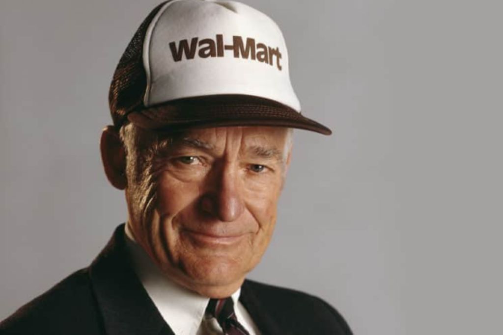 Sam Walton: Walmart's founder standing in front of a grey background staring into the camera, wearing a black business suit and a "Wal-Mart" cap