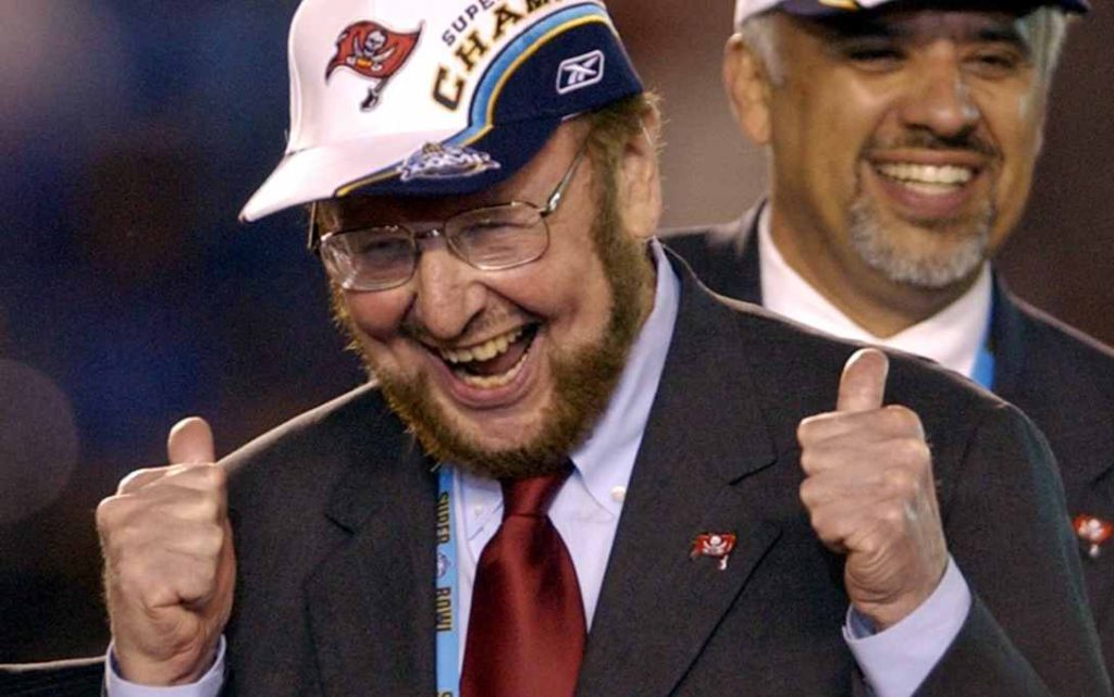 Malcolm Glazer: a man with a beard in a suit celebrating whilst wearing a Tampa Bay Buccaneers lapel badge and cap, with the head coach in the background