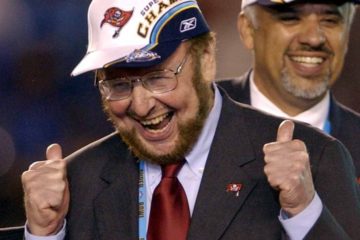Malcolm Glazer: a man with a beard in a suit celebrating whilst wearing a Tampa Bay Buccaneers lapel badge and cap, with the head coach in the background