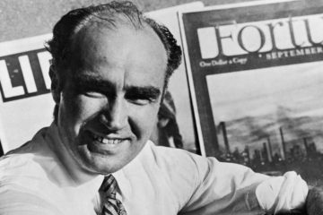 Henry Luce: a black and white photograph of a balding man sat smiling in front of some magazine covers reading "Life" and "Fortune"