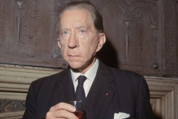 An elderly J. Paul Getty dressed in a suit holding a glass of alcohol in front of a fireplace